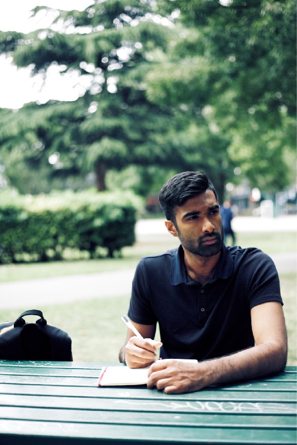 A photo shows a colored young man sitting at a desk outdoors and thinking while writing in a notebook.