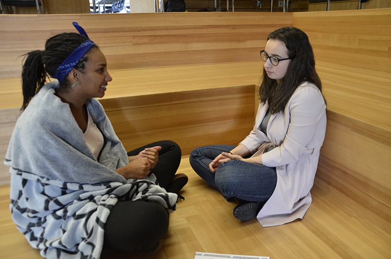 A photo shows two young women sitting on the floor of a wooden cubicle and talking to each other.