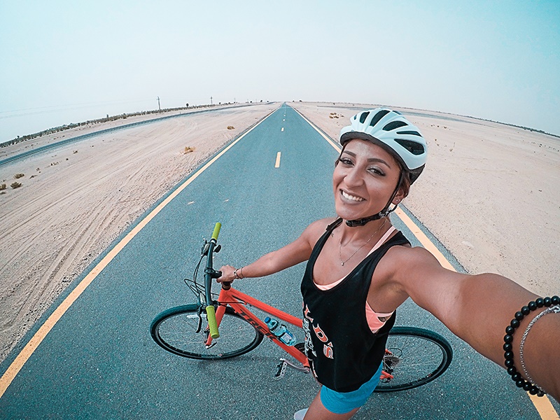 A photo shows a female cyclist in a bike helmet holding a bicycle in an empty road while smiling for a selfie.