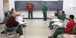 Two incarcerated men standing at a chalkboard, surrounded by other students sitting at desks.