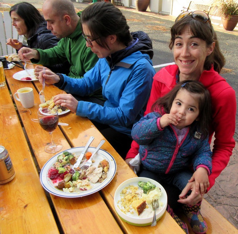 A young girl sits in her mother's lap during a holiday meal as a group of adults around the table enjoy food and drinks.