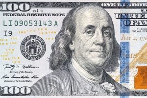 A drawing of Benjamin Franklin is seen on the face of a $100 bill. The bill is cropped to focus on Franklin's face, which is unsmiling and looking directly at viewer.
