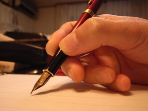 A hand is holding a red and black pen with a gold tip, poised to write on a blank piece of paper.