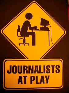 A yellow street sign has been altered to read Journalists at Play with a cartoon silhouette of a person sitting at a computer typing.