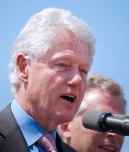 A close-up of former President Bill Clinton shows his white hair, partial profile and blue collared shirt. He is speaking into a microphone and is outside. There is a person next to him whose face is blocked by the microphone.