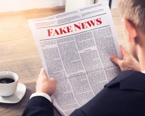 A man sits at a table near a cup of coffee reading a newspaper. His back is to the viewer. The viewer can see the headline of the newspaper that has a big, red-font headline reading, Fake News.