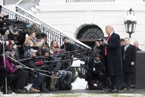 Then-President Donald J. Trump stands in a long dark coat and red tie outside a snowy White House, talking to a group of reporters huddled and crouched down and holding several long microphones.