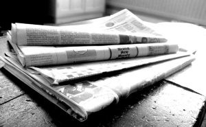 A black and white photo of four newspapers folded up and piled on top of each other on a wood table.