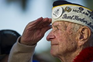 An older white man can be seen in profile, saluting and wearing a Pearl Harbor Survivor hat decorated with pins describing his military ties.