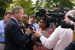 A group of people stand outside with a background of green trees. In the foreground, a man in a suit is talking to a gaggle of reporters and camera people.