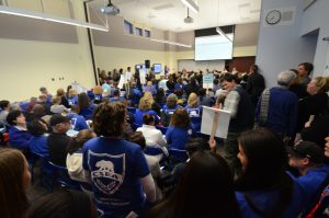 A sea of people wearing bright blue shirts can be seen facing away from the viewer toward a large white projection screen. Below that in the far distance sit members of a school board during a meeting.
