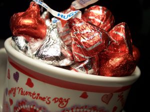 A white mug decorated with red hearts and the words, "Happy Valentine's Day" is filled with red and gold Hershey's kisses candies.