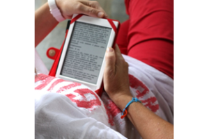 Close-up of a person seated in a relaxed pose, reading an e-book on a tablet. The individual is wearing a red shirt and holding the tablet with both hands. The screen displays pages of text, clearly indicating an e-reading session. The person has a friendship bracelet on their wrist, and their lap is covered with a white and red patterned fabric, adding a cozy and comfortable ambiance to the scene.