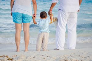 Two adults holding the hands of a tolder in front of the ocean.