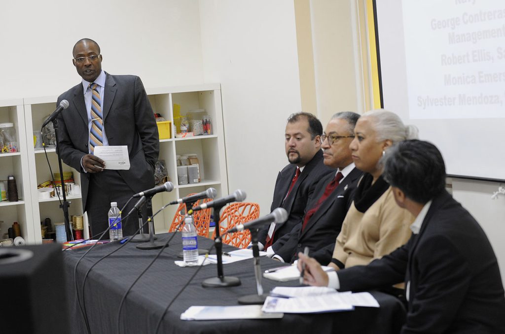 A man stands at a microphone while moderating a panel discussion of four peoiple who are seated at a table