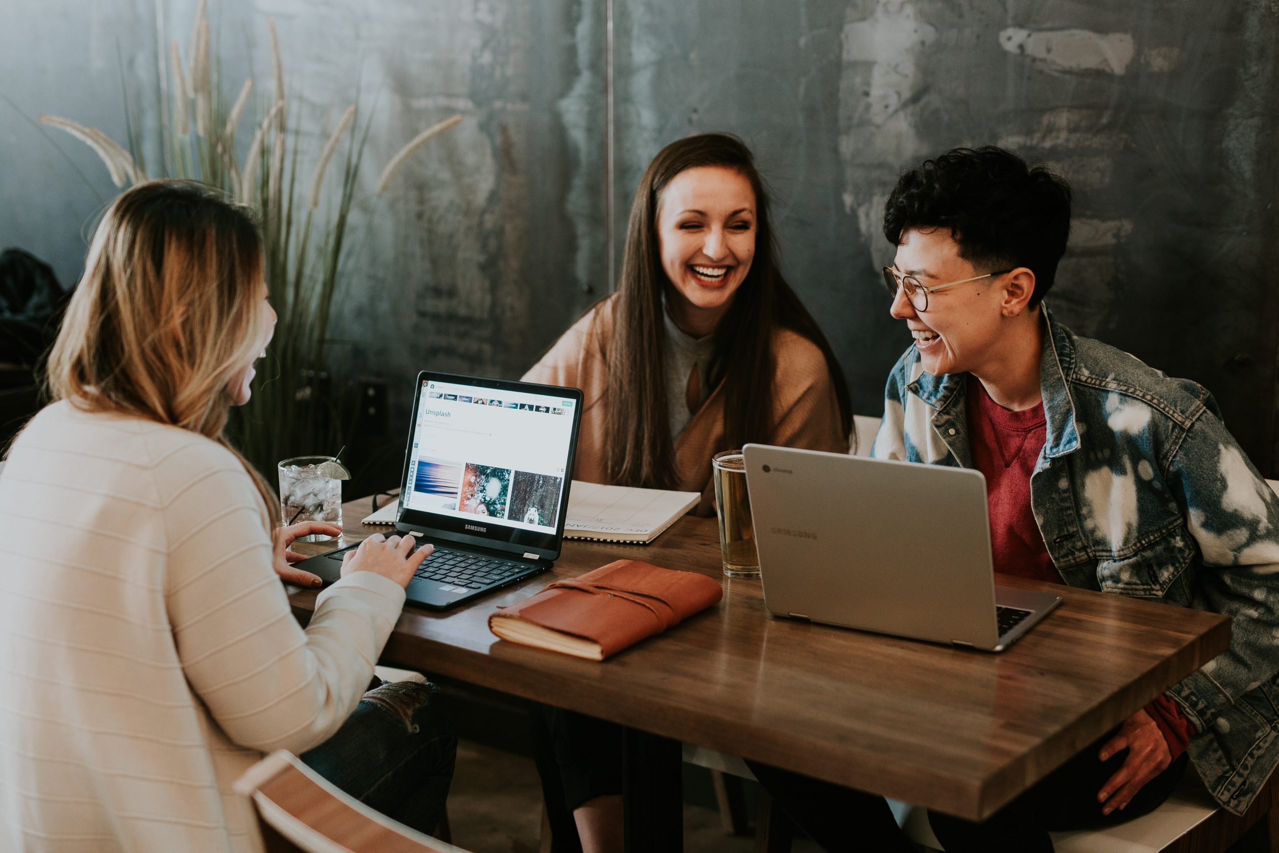 Three people sit at a table with laptops laughing