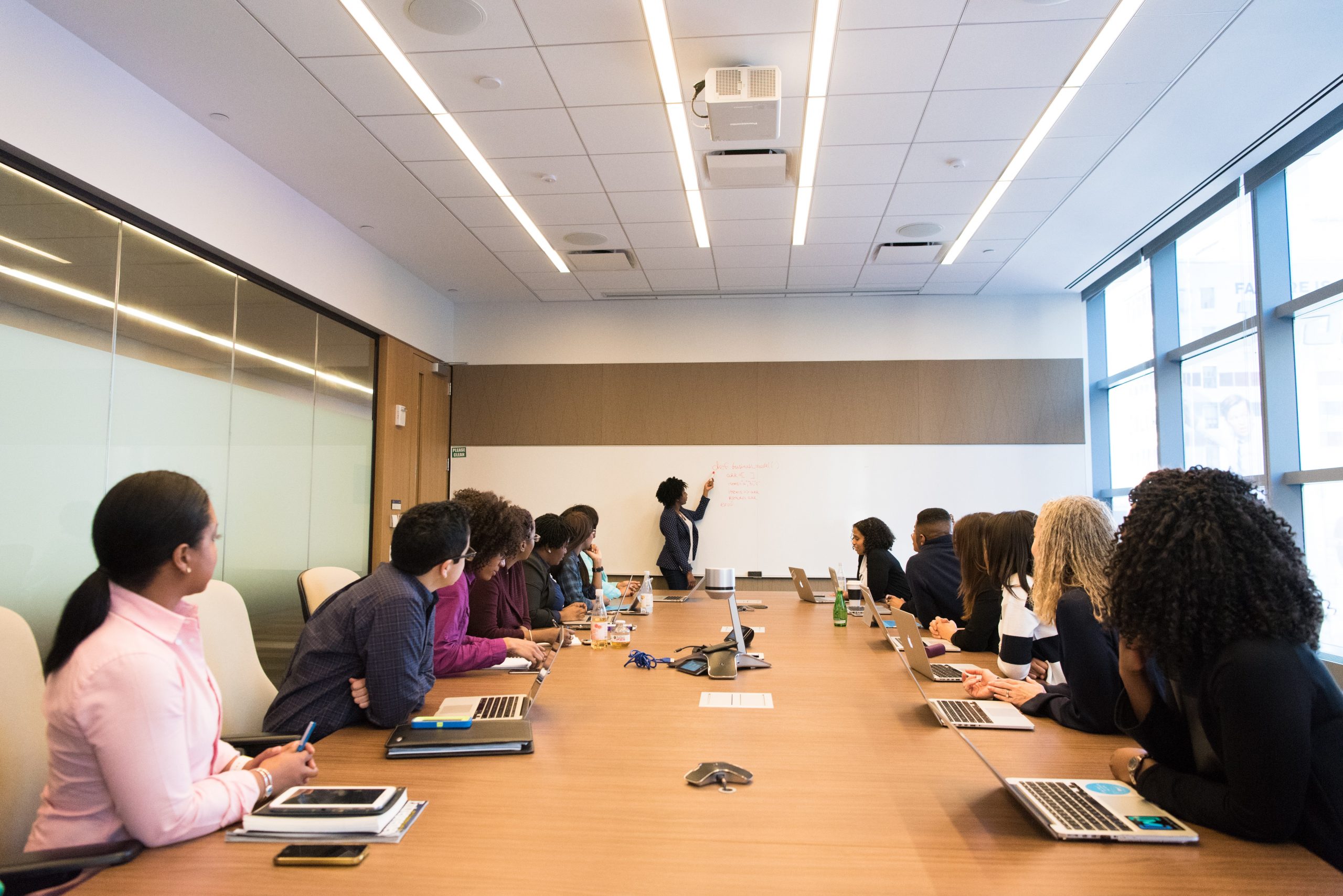 People sit at a long rectangular table with a person at a white board at the far end