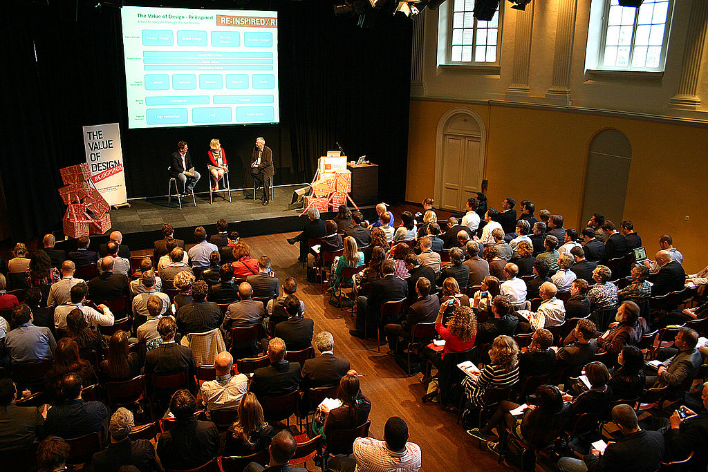 Three people sit on stools on a stage before an audience with a screen with a visual aid behind them