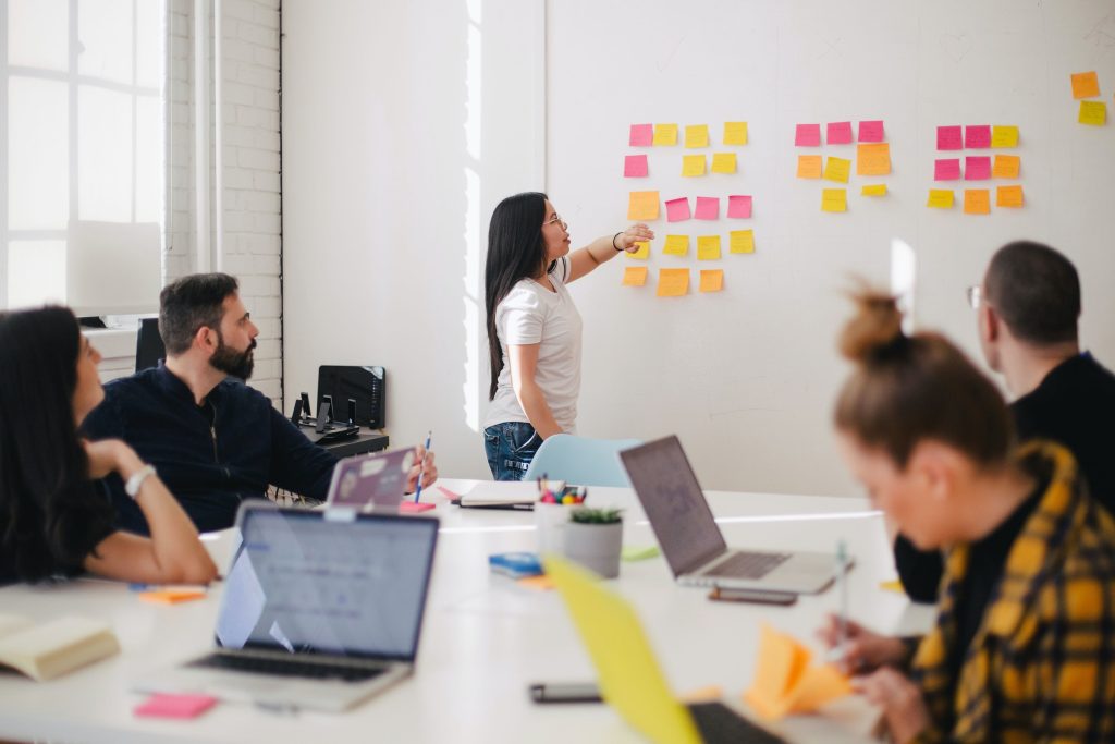 A woman points to Post-it notes organized in clusters on a wall while group members look on