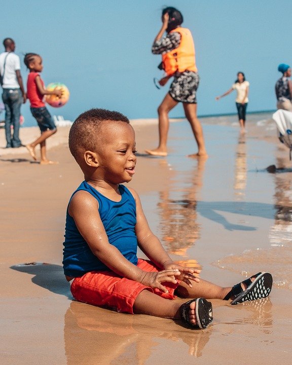 The image shows a young African boy sitting on the beach, smiling as he plays in the shallow water. In the background, there are several other people, likely tourists or beachgoers, walking along the shore or standing in the water. The boy is wearing a blue tank top and red shorts, and appears to be enjoying himself as he sits in the wet sand, the gentle waves lapping around him on a sunny day at the beach.