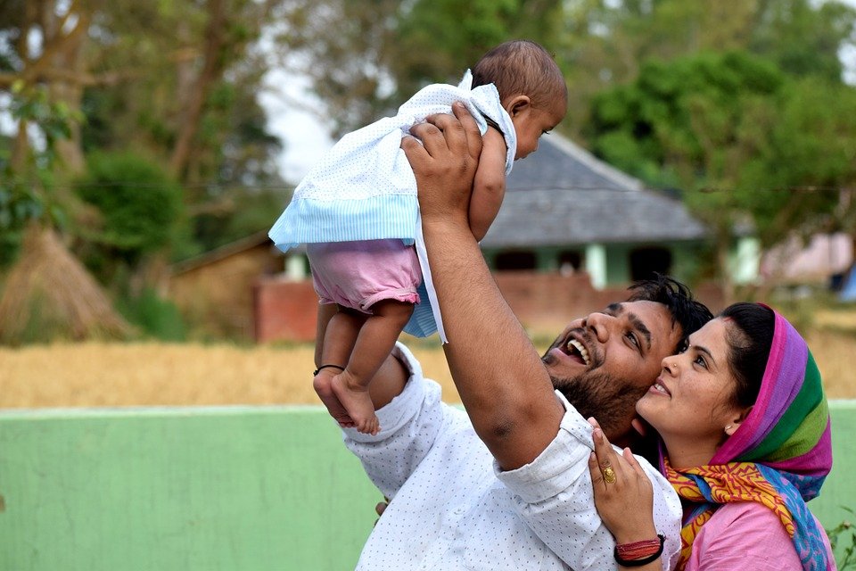 The image shows a smiling man holding up a happy baby girl outdoors in a rural setting with small buildings visible in the background. The man and baby both have cheerful expressions as he lifts her up playfully. The baby is wearing a light-colored short-sleeved outfit, while the man wears a white t-shirt. Next to them, a woman in a colorful headscarf smiles up at the baby, sharing in the joyful moment between the man and child.