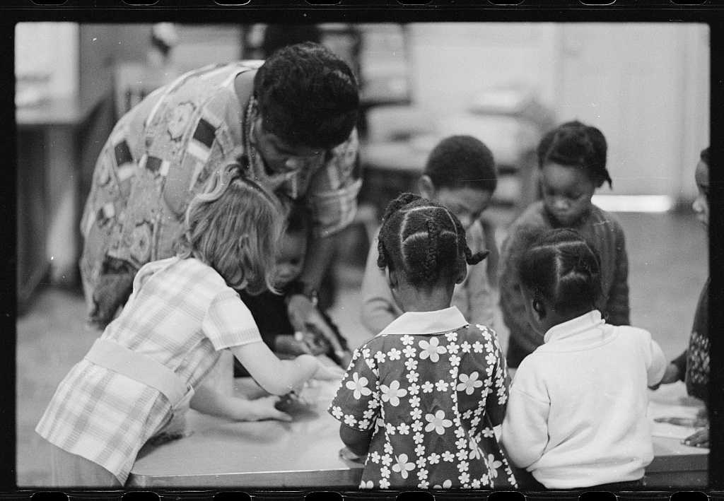 A black and white photograph showing a teacher or caregiver standing over a group of young children seated at a table. The adult appears to be assisting or instructing the children with an activity. The children have their backs to the camera and are wearing patterned shirts. The scene seems to depict a classroom or daycare setting from an earlier time period based on the clothing and hairstyles.
