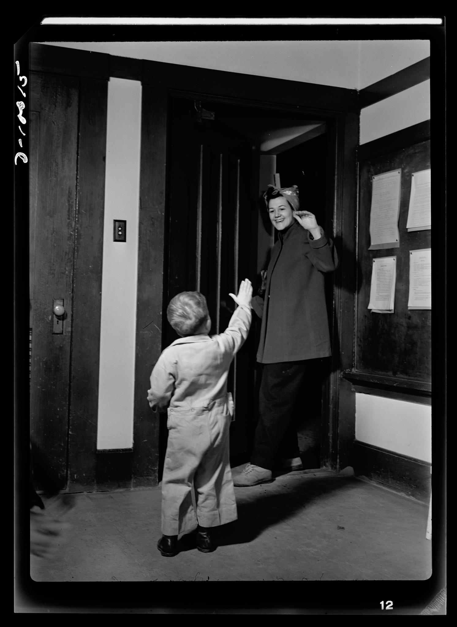 The black and white photograph shows a woman and young child, likely mother and son, standing in a doorway. The woman is smiling and has her hand raised, perhaps waving to or greeting someone outside the frame. The child is reaching up towards the woman. Based on their clothing and the setting, the image appears to be from an earlier time period, possibly the mid-20th century. A room with windows can be seen in the background through the open doorway.