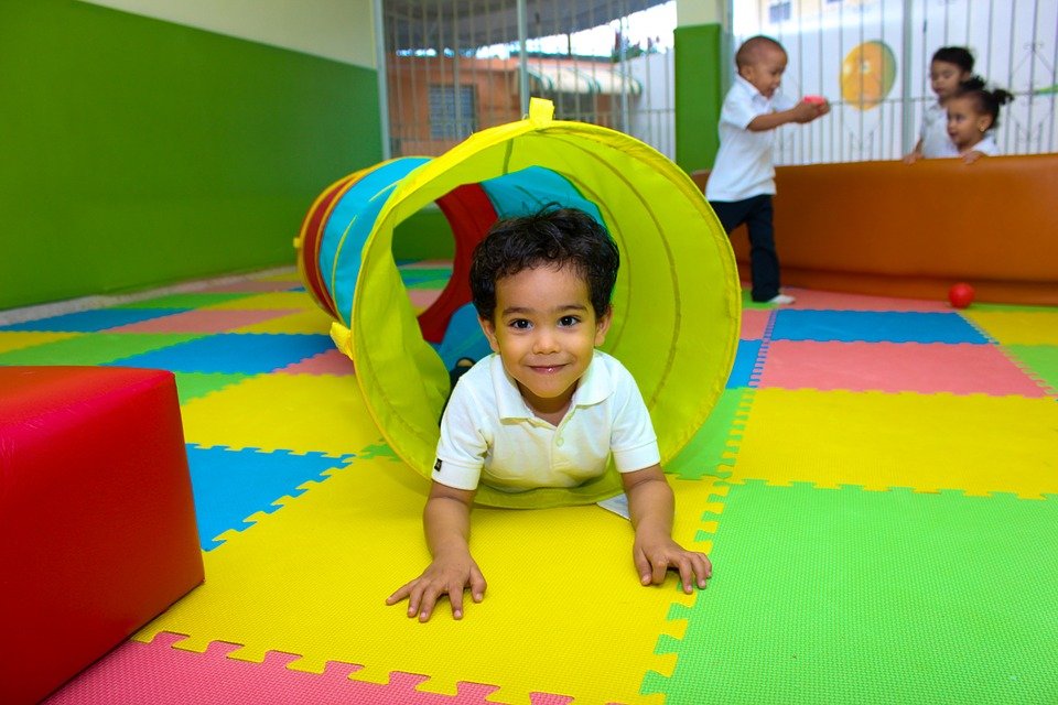The image shows a smiling young child crawling through a colorful play tunnel in what appears to be a daycare or preschool setting. Other children can be seen playing in the background. The play area has interlocking foam mats in bright colors like red, yellow and green, providing a safe and engaging space for the children to explore and have fun.