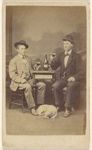 Two men drinking at a table, a dog lying at their feet], 1865–1879