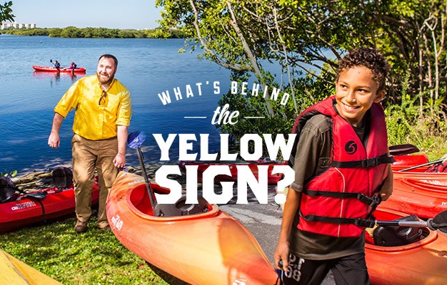 KOA advertisement asks "What's behind the yellow sign?" as two people haul a canoe