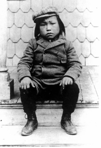 a black and white photo of a young boy in a coat and hat sitting on a step