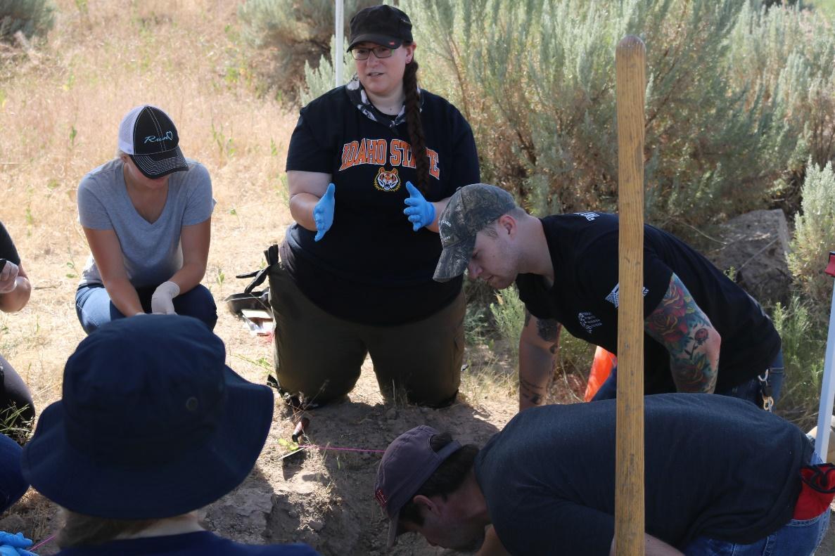 A group of individuals around an excavation. The woman in the center of the photo is talking to the rest of the group.