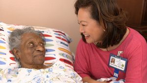 African American elder in hospital bed with Asian female RN sitting next to her