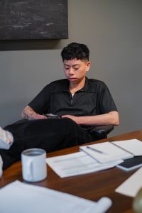 A non-binary person sitting behind a desk, using a cellphone.