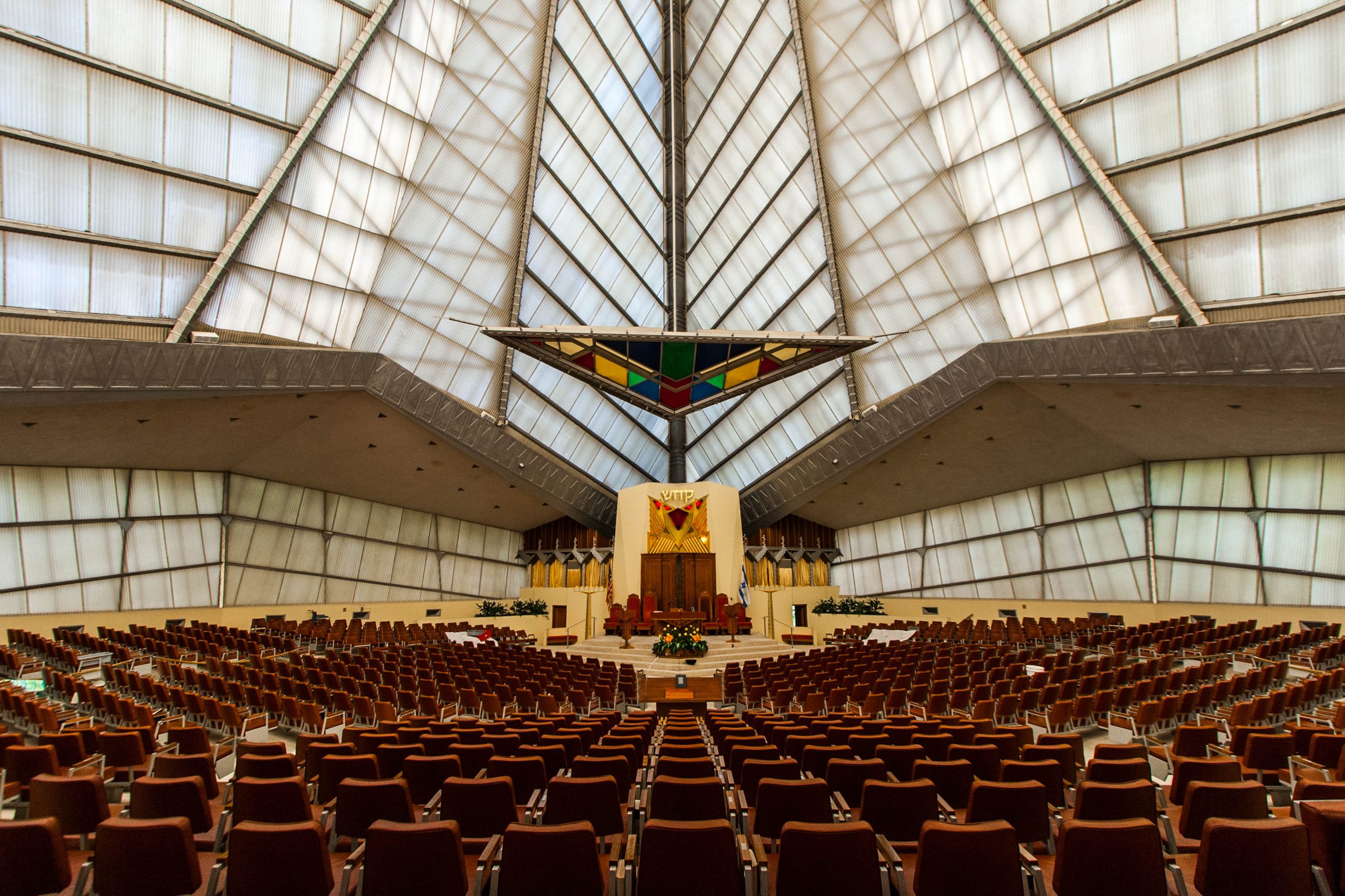 Photo of the Interior of the Beth Sholom Synagogue in Elkins Park, Pennsylvania with a very high, white windowed ceiling and colorful stained glass at the front