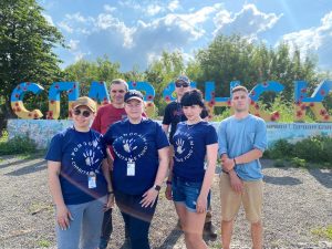 Photograph showing 6 adults, three in matching t-shirts with the words ‘Pomogayem Charitable Foundation’ upon them, in front of a large blue and yellow sign