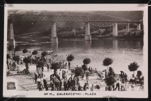 A postcard showing people walking and enjoying a promenade and beach, with a large bridge over the river, in the distance. The postcard is entitled ‘No 11 - Zeleszczyki: Plaza’