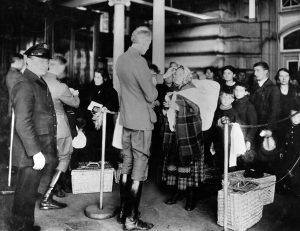 Photograph showing men in suits and shiny long boots, standing in a large hallway guarded by a man in uniform. A queue of people wearing poor, civilian clothes and carrying wicker suitcases are waiting to be examined