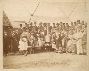 Old sepia photograph showing a number of men and women, posed under the awning of a tent, around what may be a large samovar or tea urn