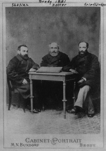 Black and white photograph of three bearded men, wearing heavy black coats, sitting around a small table. The photograph has hand written script above reading “Brody 1881” and has labelled the three men by their names: Shatiez, Netter, Friedlander