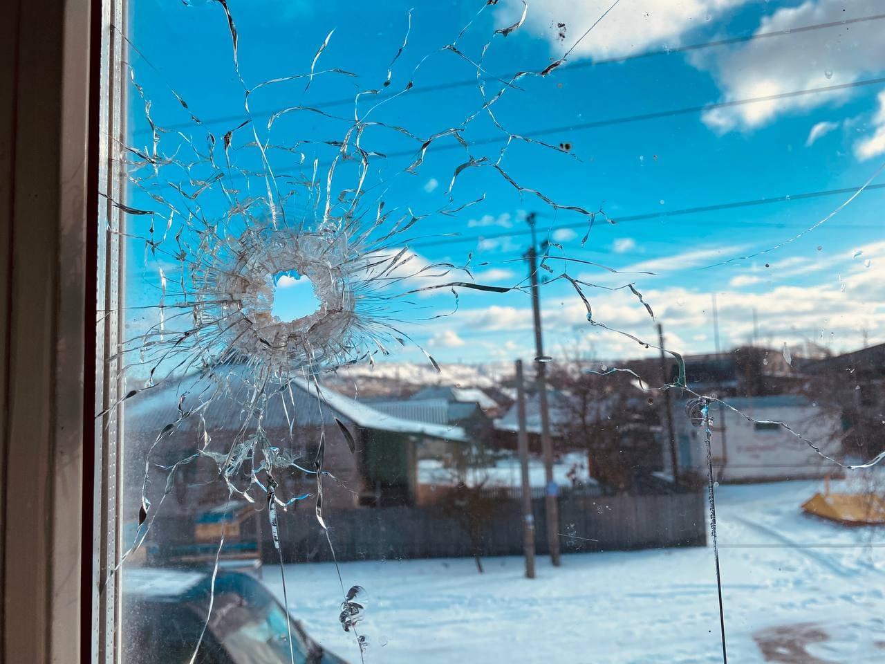 Photograph taken through a window, looking out on to a snowy landscape. The window been damaged by a bullet or shrapnel, and has a hole with cracks radiating out from it