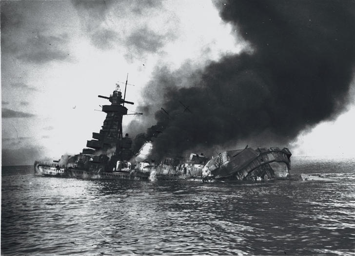 A black-and-white photo of a sinking ship that is on fire. A large plume of smoke is rising from the ship into the sky.