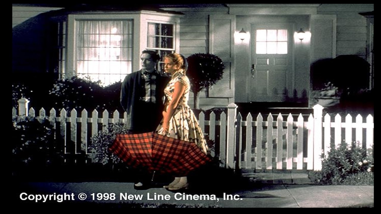 A black-and-white photo of a couple standing by a white picket fence in front of a house. The woman is in colour, wwearing a yellow 1950s style dress and a red plaid umbrella.