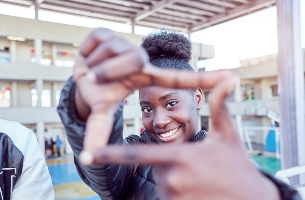 A smiling young Black woman looks through a frame made from her index fingers and thumbs held together. Une jeune femme noire souriante regarde à travers un cadre fait de ses index et de ses pouces réunis.