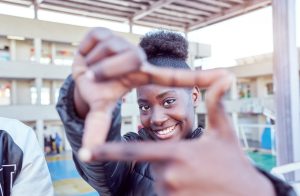 A smiling young Black woman looks through a frame made from her index fingers and thumbs held together. Une jeune femme noire souriante regarde à travers un cadre fait de ses index et de ses pouces réunis.