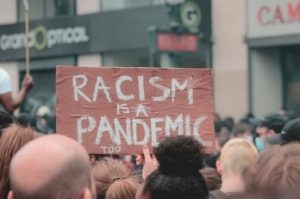 unrecognizable crowd of people with one holding a poster that says "racism is a pandemic too"