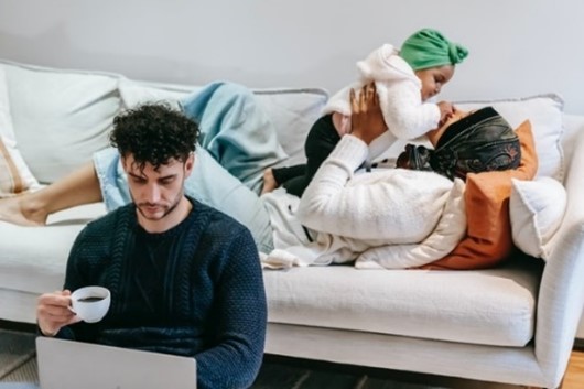 woman laying on couch and playing with a baby while man sits on the floor holding a small coffee cup and working on a laptop