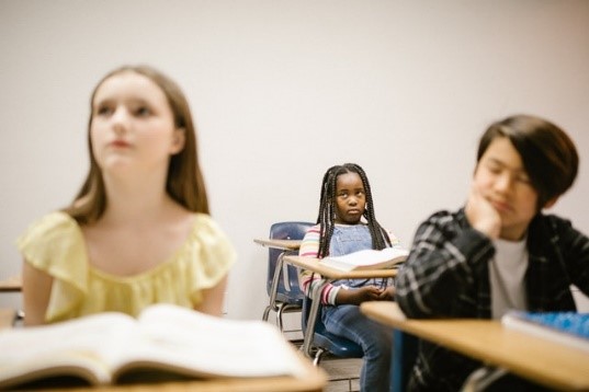 An African American girl sitting in a desk with two, out of focus other children sitting in desks in front of her
