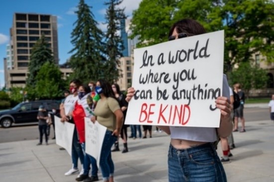 Person holding sign that says "In a world where you can be anything, be kind."