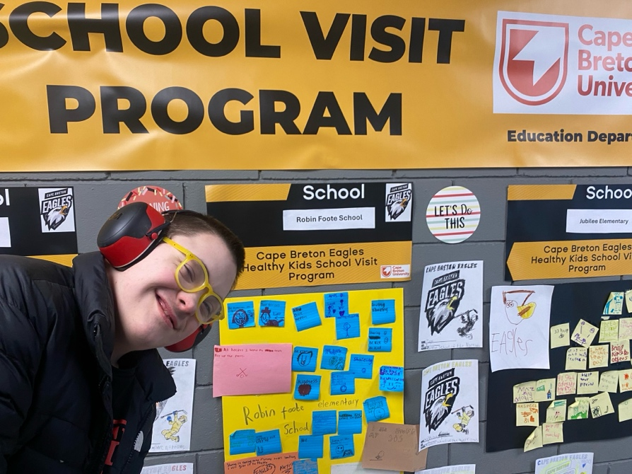 A student standing before a wall showcasing highlights of the School Visit Program.